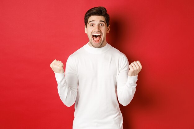 Image of happy goodlooking man in white sweater winning something making fist pump and smiling amaze...