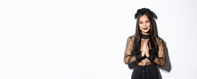 Image of happy and grateful young asian woman in gothic lace dress clasp hands together to say thank