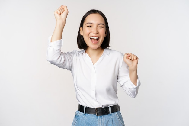 Free photo image of happy lucky asian woman hooray gesture winning and celebrating triumphing raising hadns up and laughing standing over white background