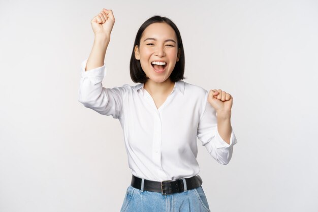 Изображение счастливой удачливой азиатской женщины, победившей и празднующей триумф, поднимающей хеднс и смеющейся, стоя на белом фоне