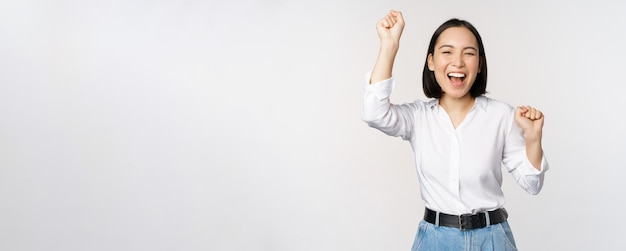Image of happy lucky asian woman hooray gesture winning and celebrating triumphing raising hadns up and laughing standing over white background