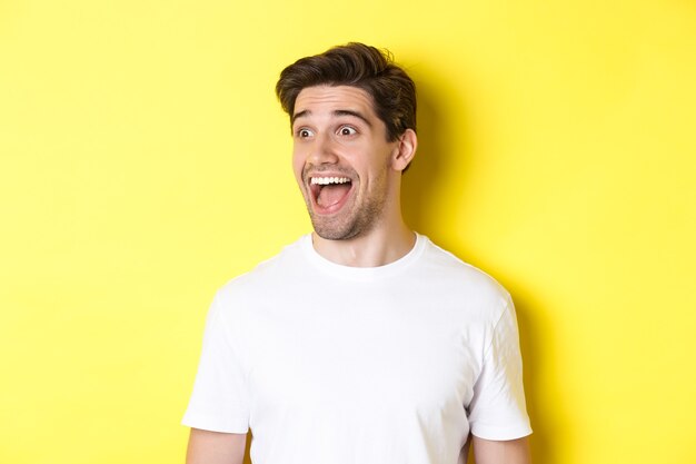 Image of happy man checking out promo, looking left with amazement, standing in white t-shirt against yellow background.