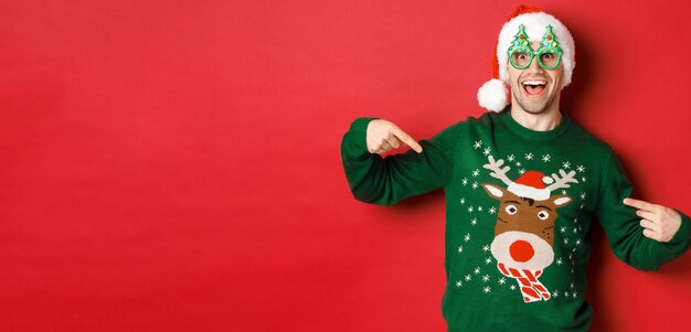 Image of happy man in party glasses and santa hat pointing at his christmas sweater and smiling stan...