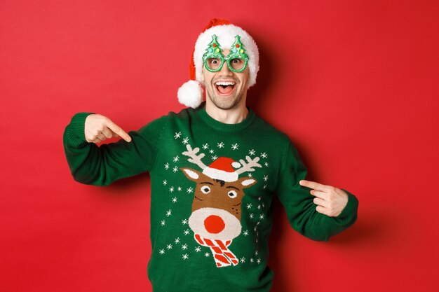 파티 안경과 산타 모자를 쓴 행복한 남자의 이미지, 크리스마스 스웨터를 가리키며 웃고, 빨간색 배경 위에 서 있는 이미지