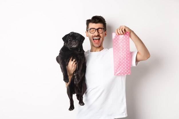 Image of happy man, pet owner, holding cute black pug and dog poop bag, standing over white background