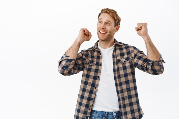 Image of happy redhead man celebrating sport victory, dancing and looking up as if watching sports game on screen above, standing positive against white background