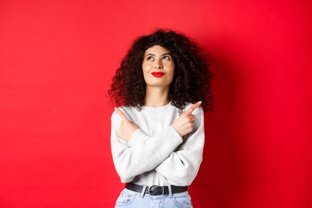 Image of happy smiling woman pointing sideways, looking up with determined face, choose something, standing confident in her decision on red background