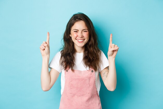 Image of happy smiling young woman with curly hair, laughing and pointing fingers up at logo banner, showing promo deal, standing against blue background.