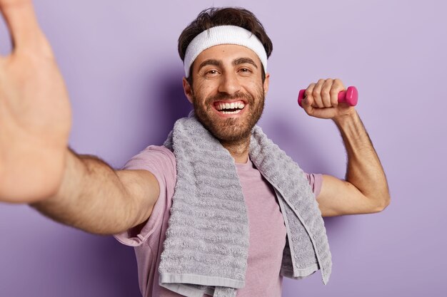 Image of happy sportsman has workout indoor, makes selfie with dumbbell