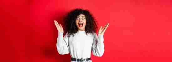 Free photo image of happy and surprised curly woman in makeup and sweatshirt raising hands up and rejoicing fro