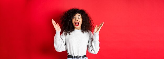 Image of happy and surprised curly woman in makeup and sweatshirt raising hands up and rejoicing fro