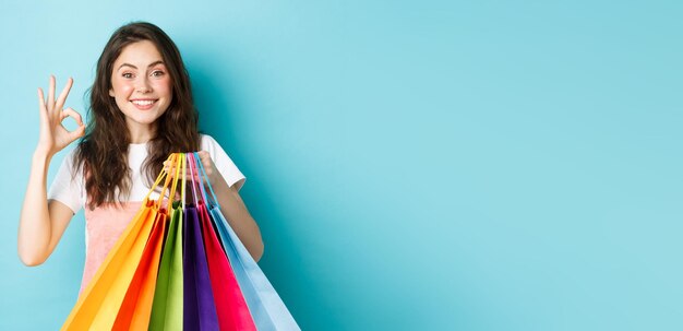 Изображение счастливой молодой гламурной женщины, делающей покупки в магазинах со скидками, показывающей знак "хорошо"