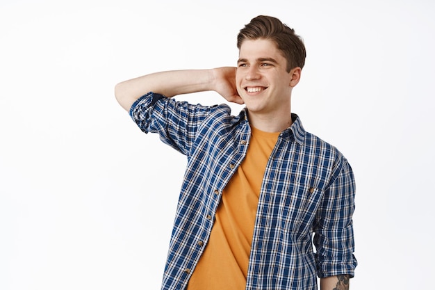 Image of happy young man smiling, looking aside carefree, touching hair and look pleased at logo, standing relaxed in casual outfit against white background.