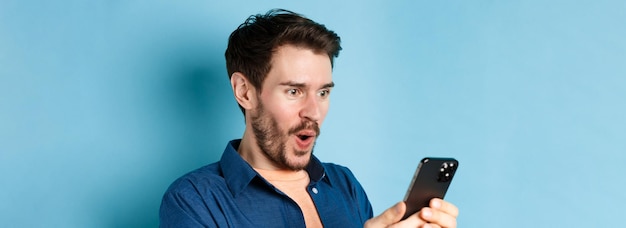 Free photo image of happy young man stare at mobile screen winning online and cheering standing on blue backgro