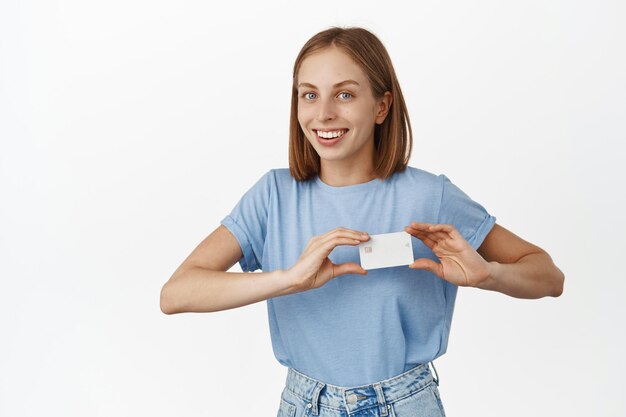 Image of happy young woman showing discount, credit card near heart, likes bank new offer, cashback or deposit proposal, smiling satisfied, white background