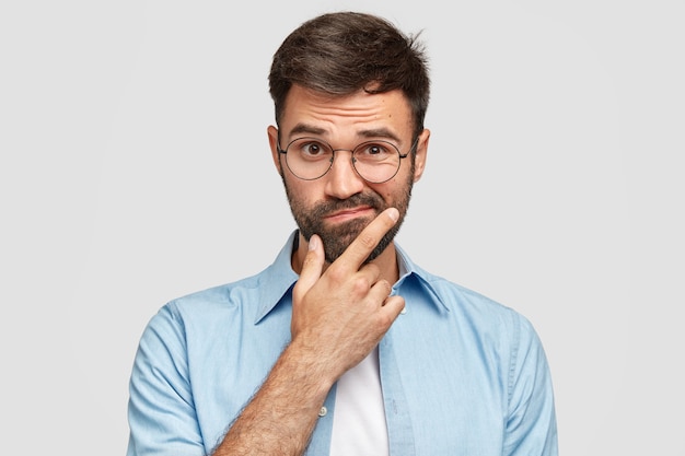 Image of hesitant unshaven European man with thick beard, holds chin, purses lips with clueless expressions