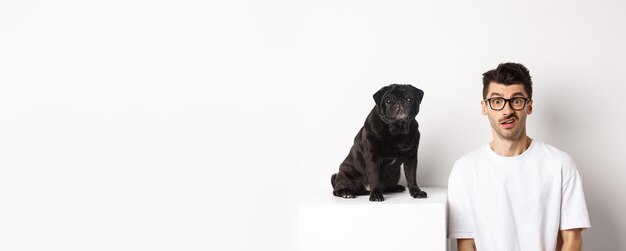 Free photo image of hipster dog owner sitting near cute black pug staring at camera confused and puzzled white