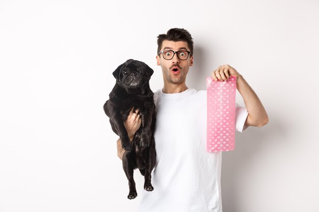 Image of hipster guy pet owner, holding cute black pug and dog poop bag, standing over white background.