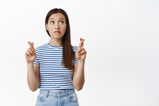 Image of hopeful 20s girl holds fingers crossed, bites lip and looks nervous up, waiting for results, praying, making wish for dream come true, standing against white background