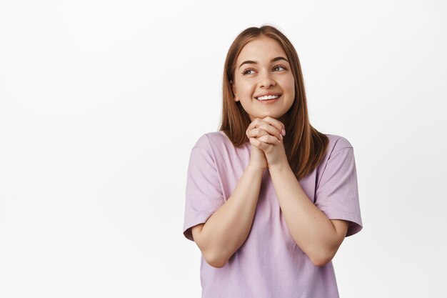 Image of hopeful smiling blond woman, looking away thoughtful, clench hands, thinking of something, dreaming or imaging beautiful things, standing against white background.