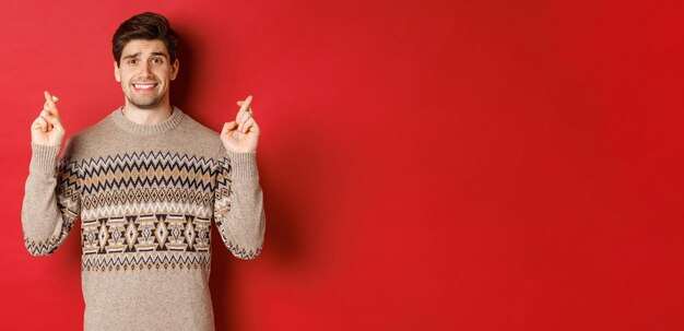 크리스마스 스웨터를 입은 희망적이고 걱정스러운 남자의 이미지는 행운을 위해 손가락을 교차하고 새해 선물 빨간색 배경에 대해 불안하게 만드는 무언가를 기다리고 있습니다.