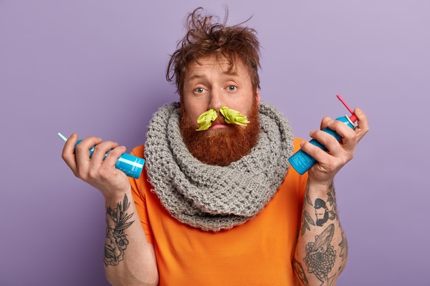 Free photo image of ill red haired man has running nose, handkerchief in nostrils, wears knitted warm scarf on neck