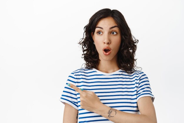 印象的なブルネットの女の子のあえぎ、素晴らしいディスカウントショップを指差して左を見て、コピースペースにロゴを表示し、白い背景の上にストライプのTシャツで立っている画像。