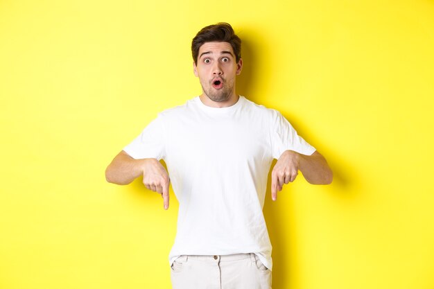 Image of impressed guy pointing fingers down, gasping amazed and looking at camera, standing over yellow background.