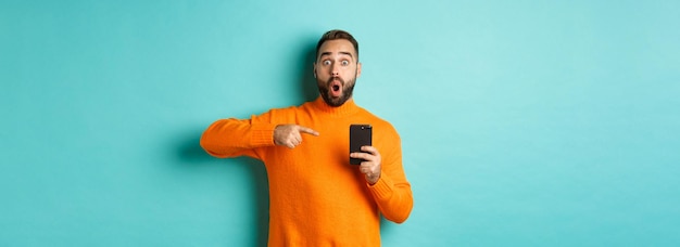 Image of impressed man showing something on mobile phone pointing at smartphone and gasping amazed s