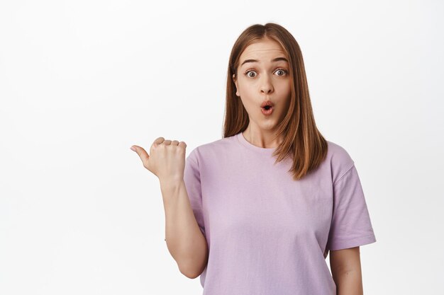 Image of impressed natural girl gasp, look astonished by promo deal, event advertisement, pointing finger left and say wow astonished,s tanding against white background