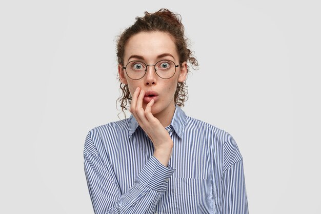Image of impressed stunned freckled female hears surprising news, keeps hand on cheek, dressed in striped shirt, stands over white wall. Astonished amazed young woman with bated breath