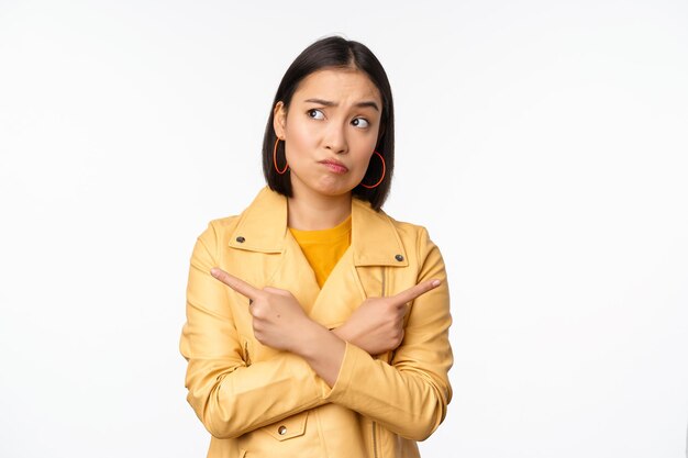 Image of indecisive asian girl pointing fingers sideways pointing left and right choosing variant deciding standing over white background