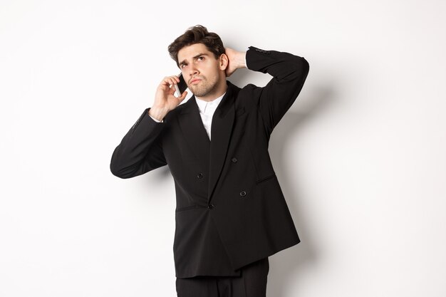 Image of indecisive businessman talking on phone and thinking, looking doubtful, making decision, standing over white background