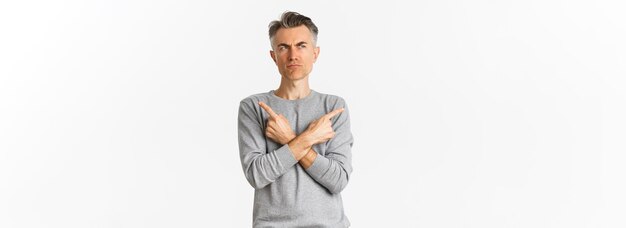 Image of indecisive middleaged man making choice pointing fingers sideways and frowning doubtful standing unsure over white background