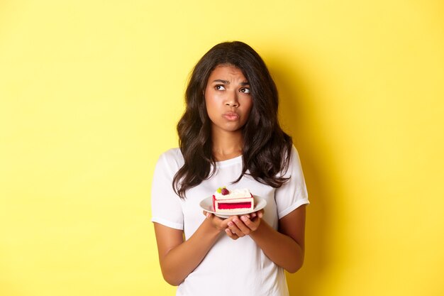 Image of indecisive and sad african-american woman, looking upset, cannot eat cake, standing over yellow background