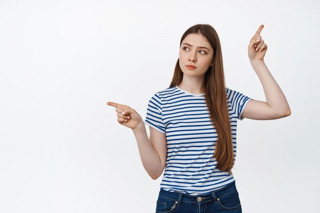 Image of indecisive teenage girl pointing sideways with thoughtful face, making decision, standing on white.