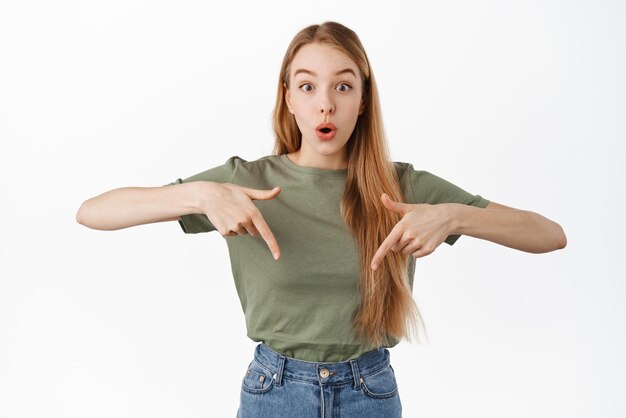 Image of intrigued blond girl gasp say wow and points fingers down at interesting advertising promo offer asks question about product stands over white background