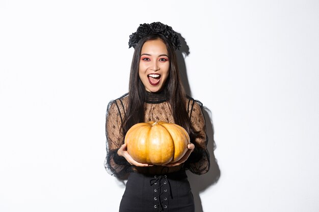 Image of joyful asian woman in witch costume, celebrating halloween, giving you pumpkin and looking scary, standing over white background.