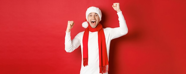 Image of joyful caucasian man in santa hat and scarf, shouting for joy and raising hands, celebrating victory or win, triumphing over red background