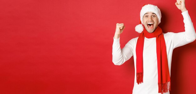 サンタの帽子とスカーフを身に着けて、喜びを叫び、手を挙げ、勝利または勝利を祝い、赤い背景に勝利した、うれしそうな白人男性の画像。
