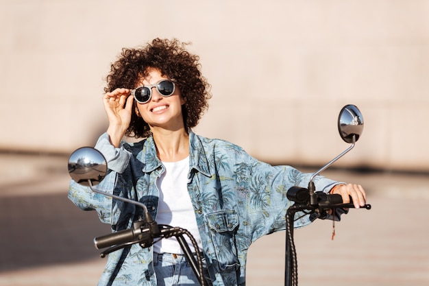 屋外のモダンなバイクに座っているサングラスでうれしそうな巻き毛の女性のイメージ