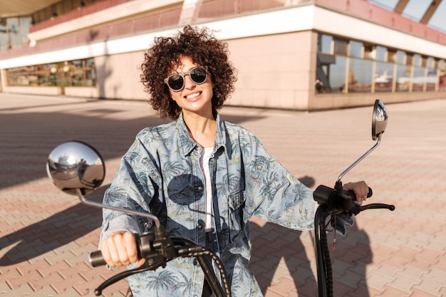 Immagine della donna riccia allegra in occhiali da sole che si siedono sulla motocicletta