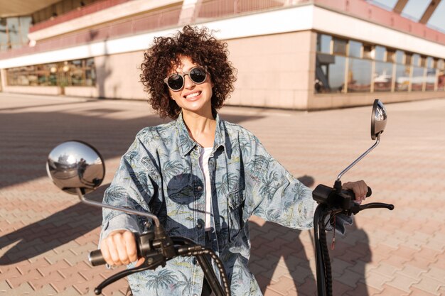 Image of joyful curly woman in sunglasses sitting on motorbike