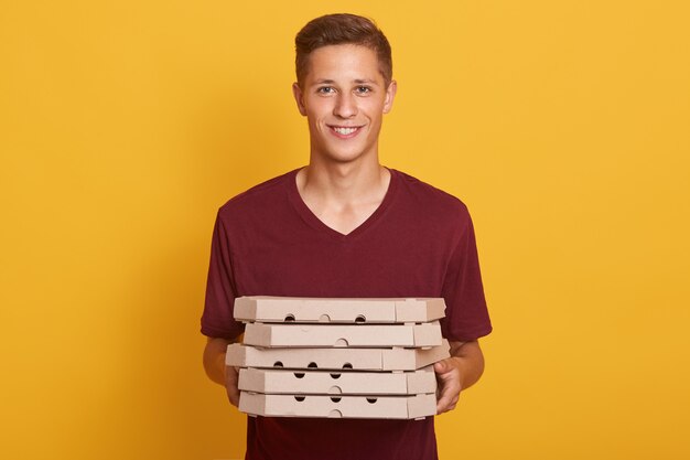 Image of joyful handsome delivery man wearing casual burgundy t shirt, holding stack of pizza boxes in hands and looking directly at camera isolated on yellow studio. Junk food concept.