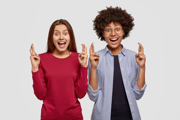 Image of joyful multiethnic women keep hands raised with crossed fingers