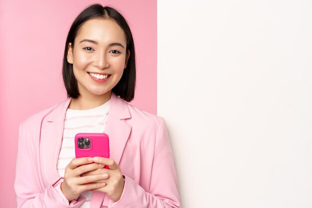 Image of korean female entrepreneur in suit standing near info wall advertisement on board holding smartphone and smiling posing over pink background