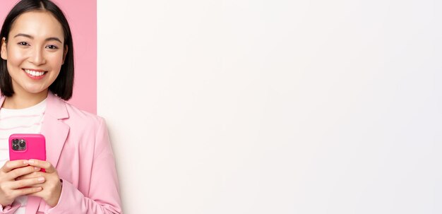 Image of korean female entrepreneur in suit standing near info wall advertisement on board holding smartphone and smiling posing over pink background