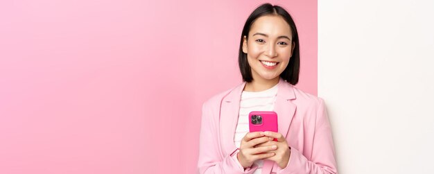 Image of korean female entrepreneur in suit standing near info wall advertisement on board holding smartphone and smiling posing over pink background