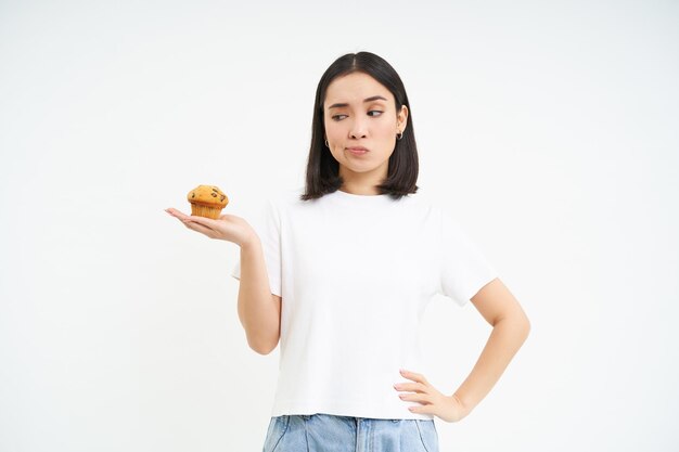 Image of korean girl looking at cupcake and thinking making decision white background