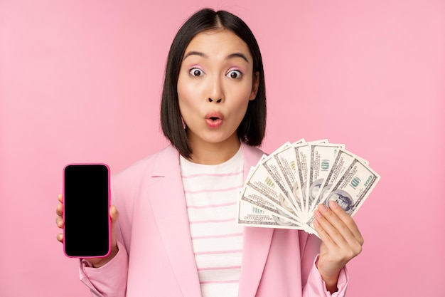 Image of korean successful corporate woman showing money dollars and smartphone app screen interface of mobile phone application concept of investment and finance pink background